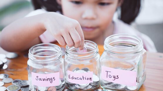 A child drops a coin into one of three coin jars labeled "Education." The other two jars are labeled "Savings" and "Toys."