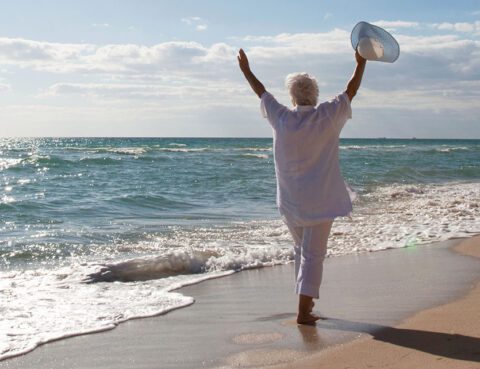 Senior woman in white walking along the beach with arms raised, symbolizing freedom and a fresh start.
