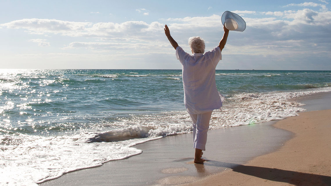Senior woman in white walking along the beach with arms raised, symbolizing freedom and a fresh start.