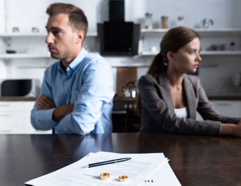 Divorcing couple sitting apart at a table with divorce papers and wedding rings in focus.