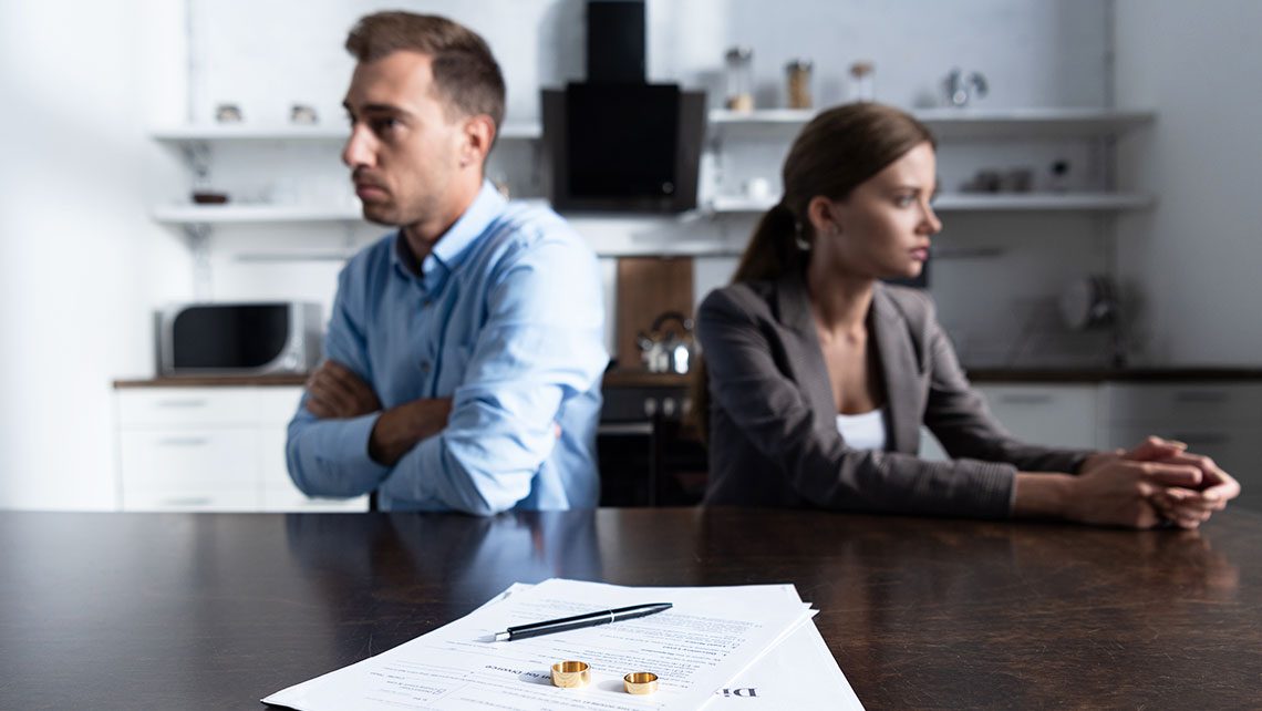 Divorcing couple sitting apart at a table with divorce papers and wedding rings in focus.