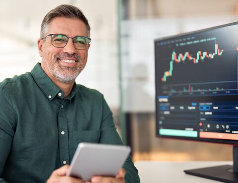 Man smiling at desk with stock trading data on screen.