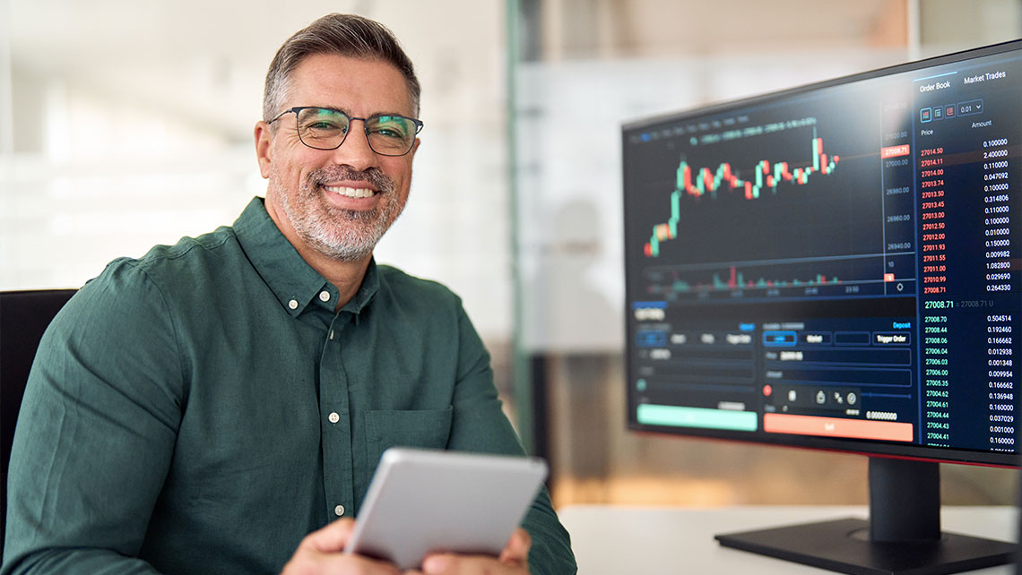 Man smiling at desk with stock trading data on screen.